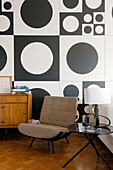 Side table and upholstered chair in front of black and white wall decoration in 60s style