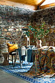 Various rugs, vase with leafy branches and candle holder on table in front of rustic natural stone wall