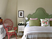 Double bed with green headboard and pretty rattan armchair in bedroom with patterned wallpaper