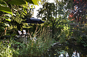 Teich im üppig bewachsenen Garten, im Hintergrund Terrassenplatz mit Sonnenschirm