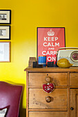 Yellow wall with Keep Calm and Carry On poster above antique wooden chest of drawers with decorative items