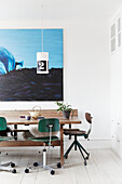 Wooden table with green desk chairs and pendant light in front of a modern painting