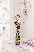 Flower arrangement in glass bottle on white wooden tray, wall with bird wallpaper
