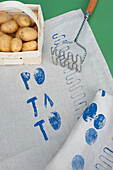 Korb mit rohen Kartoffeln, Kartoffelstampfer und blau bedrucktem Stoff
