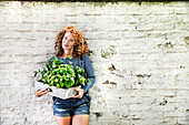 Portrait of young woman with fresh herbs in a box leaning against white brick wall