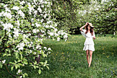 Young woman wearing white dress and floppy hat walking barefoot in garden with blossoming apple trees