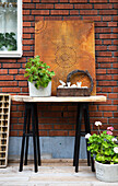 Ornaments and geraniums on and around old table on wooden terrace against brick wall