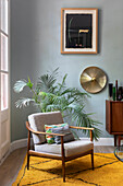 Mid-century armchair, potted palm, brass wall lamp and modern artwork on wall
