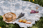 Strohhut, Lavendelblüte, Holzrett mit Birnen, Glas Wein und Buch auf Picknickdecke
