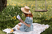 Female in dress and straw hat reading novel while sitting on picnic blanket on green meadow near swings in summer countryside