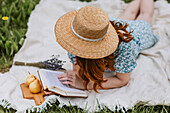 Female in dress with straw hat on face lying with open book on picnic blanket while chilling alone and enjoying summer weekend in countryside