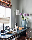 Schreibtisch mit Büchern, Tischlampe und Vase mit Alliumblüten vor Fenster