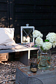 Holzsitzbank mit Laterne und Hortensienstrauß (Hydrangea) auf Metallkiste im Garten