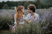Serene couple sitting in lavender field with blossoming flowers and looking at each other