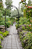 Gepflasterter Gartenweg mit Rosenbogen und umgebenden Beeten