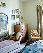Details of a traditional bedroom with a large framed mirror behind a small seat and next to a wooden chest of drawers below a number of framed images
