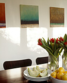 Details of a dining table laden with fruit and flowers in front of three paintings mounted on a white wall