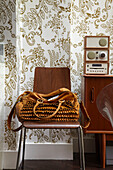 Handbag on wooden chair with stereo on sideboard and gold patterned wallpaper in home of London fashion designer