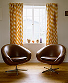 Pair of brown leather retro style swivel chairs in front of window with orange pattern curtains