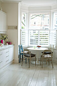 A modern kitchen with a white painted floor and a round kitchen table underneath a large bay window with shutters