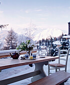 An outdoor detail of a wooden table and bench seat set on a snow covered terrace with view of mountains in the distance