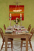 A detail of a country green dining room with a wooden table laid for Christmas lit candles table setting spotty napkins red artwork