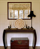 An antique wooden table with an antique wooden chest below framed map above and decorated by an antique plate candles and lamp