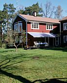 Exterior view of the back of a traditional wooden house with a awning over a table and chairs facing a large lawn
