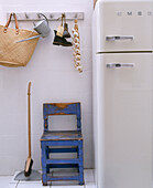 Rustikaler blauer Stuhl neben Retro-Kühlschrank darüber Korbtasche, Gießkanne, Schuhe und Knoblauchzopf an Hakenleiste hängend