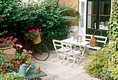 A patio area outside the back door with a white garden table and chairs next to a bike and a small table of flowers