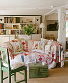 A country sitting room upholstered sofa with patchwork cover, wooden chest, painted char shelving, lamp, flower arrangement