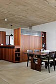 Dark wood chairs set at table on polished cement floor in open plan dining area with freestanding kitchen units