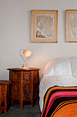 Lit lamp on carved bedside table with striped bed cover below cartographic artwork