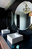 Doppelwaschbecken unter runden Spiegeln in einem schwarz getäfelten Badezimmer mit Kronleuchter