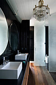Doppelwaschbecken unter runden Spiegeln in einem schwarz getäfelten Badezimmer mit Kronleuchter