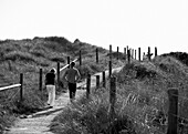 Rear view of couple walking on boardwalk in sand dunes at approach to beach