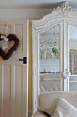 Herzförmiger Kranz an der Tür neben einem weiß gestrichenen, geretteten Kleiderschrank, der ein ganz weißes Wohnzimmer widerspiegelt