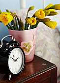 Black alarm clock and vase of daffodils on wooden bedside table