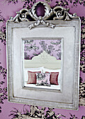Bed and cushions reflected in silver mirror hanging on patterned wallpaper