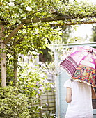 Woman shaded with colourful parasol below pergola with climbing rose in London garden UK