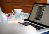 Woman sitting with teacup and saucer and laptop computer in Hastings home, East Sussex, UK