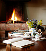 Aufgeschlagenes Buch mit Teetasse und Krug auf einem Tisch mit beleuchtetem Feuer in einem Bauernhaus in Oxfordshire, England, UK