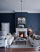 Pair of sofas with grey ottoman and lit fire in blue East Grinstead living room, West Sussex, UK