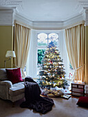 Christmas presents under tree in bay window with armchair and blanket in, UK home