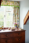 Trinkets on wooden chest of drawers at window with floral curtains in Syresham home, Northamptonshire, UK
