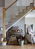 Armchair and log baskets under stairs with glass banister in Northumberland farmhouse, UK