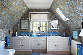 Double basins at window with blue and gold patterned wallpaper in Cotswolds bathroom, UK