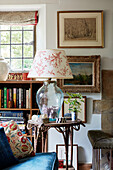 Glass lamp on side table with framed pictures and bookcase in Oxfordshire farmhouse, UK