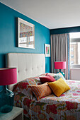 Assorted cushions and tropical cover on bed with pink lamps in London apartment, UK