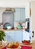 Tomatoes and bread on wooden counter with light blue fitted kitchen in, UK cottage